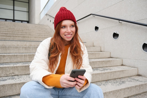 Redhead girl in red hat sits on stairs and uses mobile phone modern woman holding smartphone texting