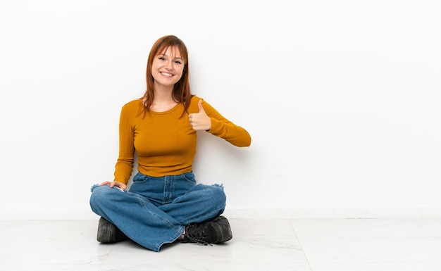 Photo redhead girl sitting on the floor isolated on white background giving a thumbs up gesture