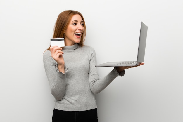 Redhead girl over white wall with laptop and credit card