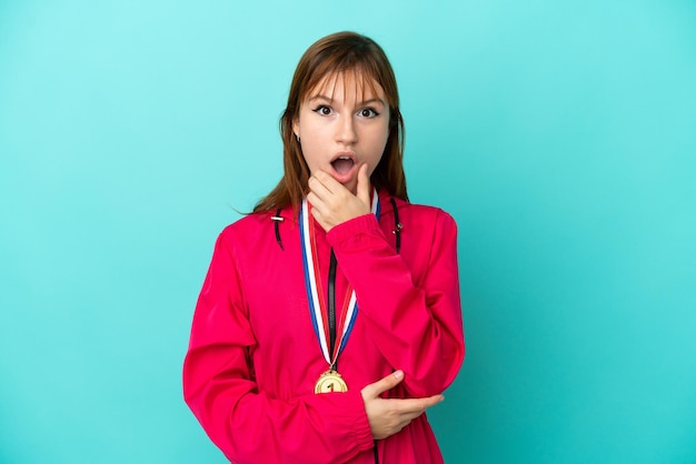 Redhead girl with medals isolated o blue background surprised and shocked while looking right