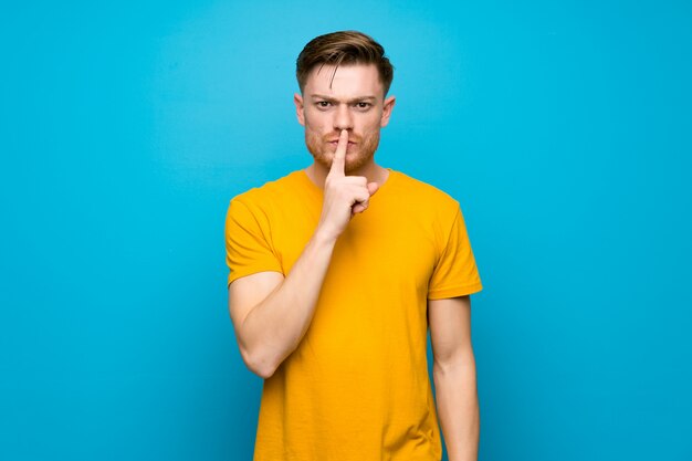 Redhead man over blue wall showing a sign of silence gesture putting finger in mouth