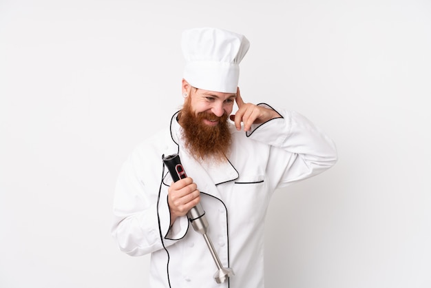 Redhead man using hand blender over isolated white wall laughing