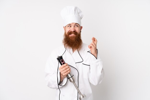 Redhead man using hand blender over isolated white wall with fingers crossing