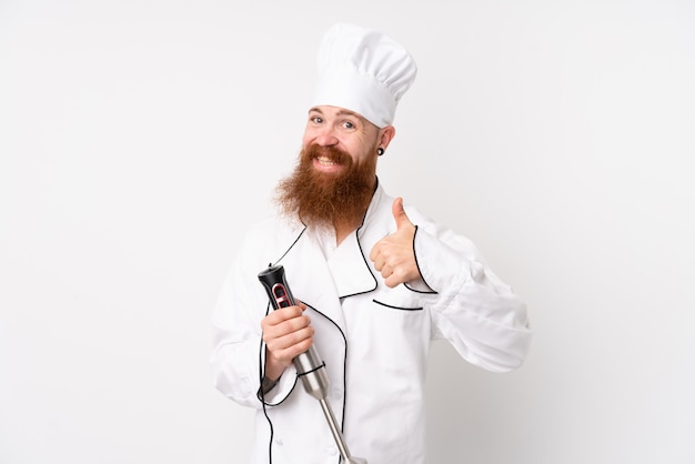 Redhead man using hand blender over white wall giving a thumbs up gesture