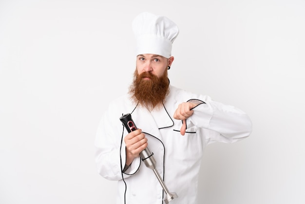 Redhead man using hand blender over white wall showing thumb down sign