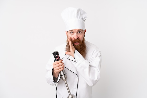 Redhead man using hand blender over white wall whispering something