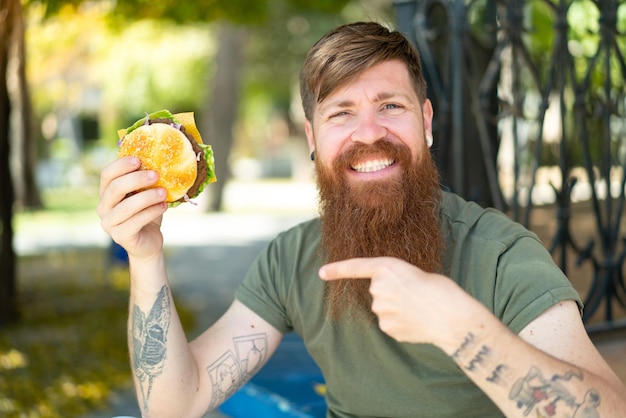 Redhead man with beard holding a burger at outdoors and pointing it