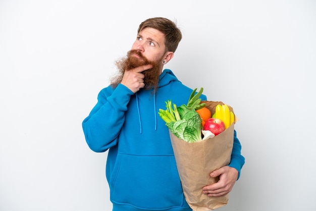 Redhead man with beard holding a grocery shopping bag isolated on white background having doubts