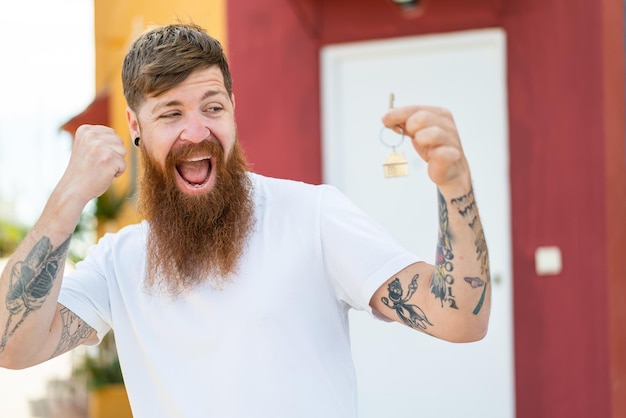 Redhead man with beard holding home keys at outdoors celebrating a victory