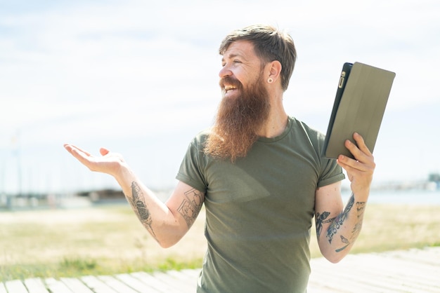 Redhead man with beard holding a tablet at outdoors with surprise facial expression