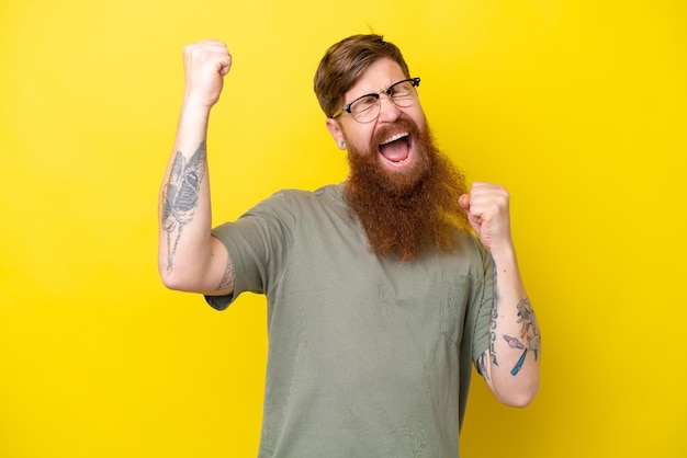 Redhead man with beard isolated on yellow background celebrating a victory