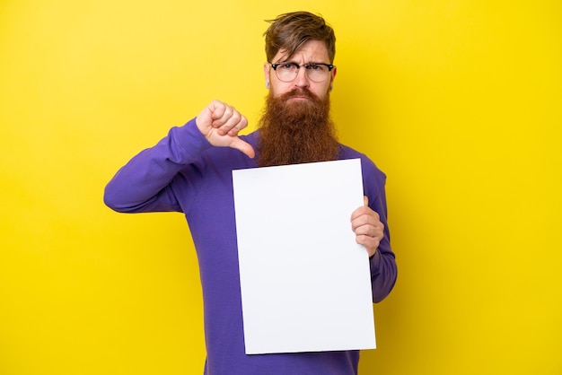 Redhead man with beard isolated on yellow background holding an empty placard and doing bad signal