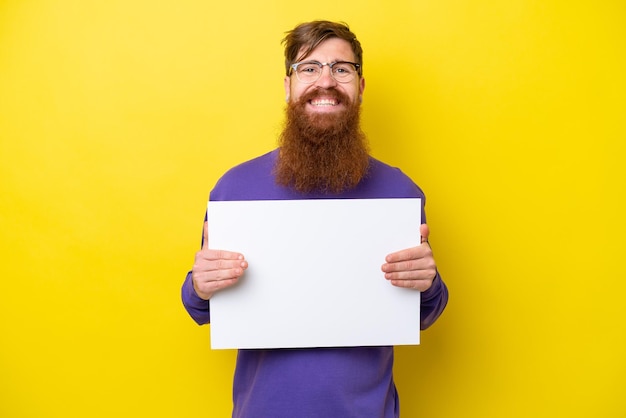 Redhead man with beard isolated on yellow background holding an empty placard with happy expression