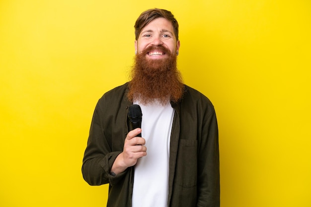 Redhead man with beard picking up a microphone isolated on yellow background smiling a lot