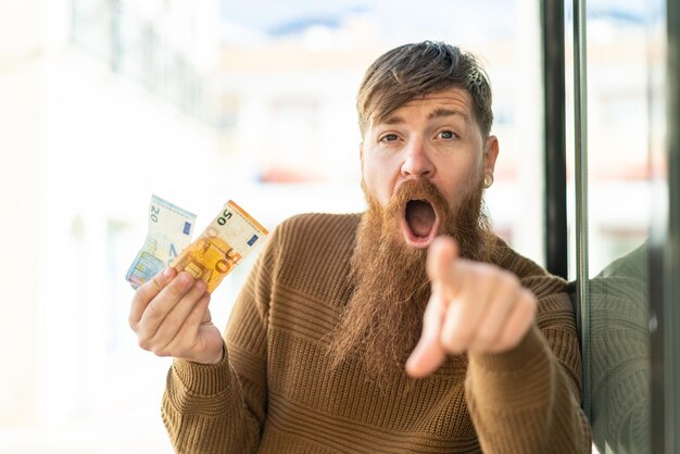 Redhead man with beard taking a lot of money at outdoors surprised and pointing front