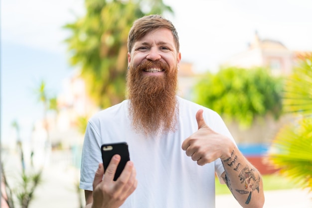 Redhead man with beard using mobile phone while doing thumbs up