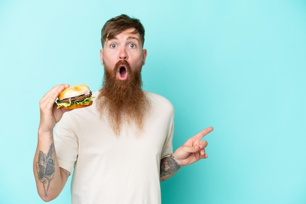 Redhead man with long beard holding a burger isolated on blue background surprised and pointing side