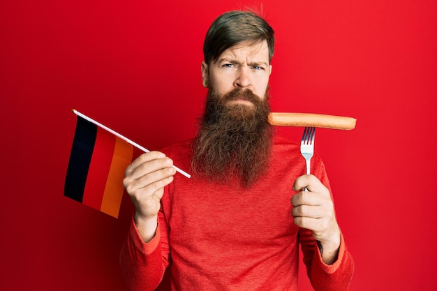 Redhead man with long beard holding fork with pork sausage and germany flag skeptic and nervous frowning upset because of problem negative person