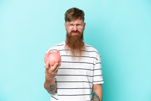 Redhead man with long beard holding a piggybank isolated on blue background with sad expression