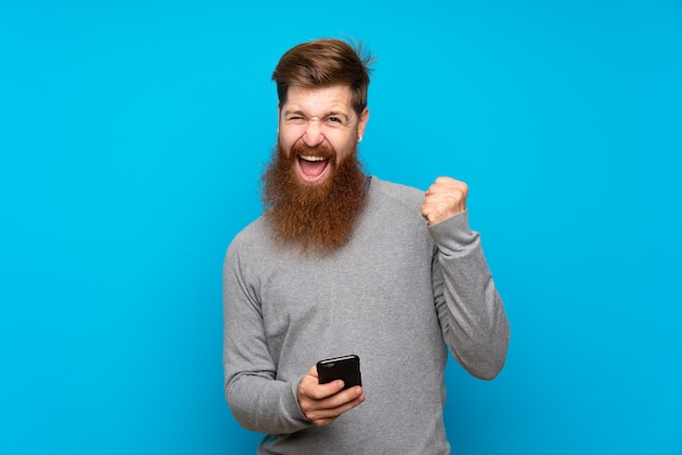 Redhead man with long beard over isolated blue with phone in victory position