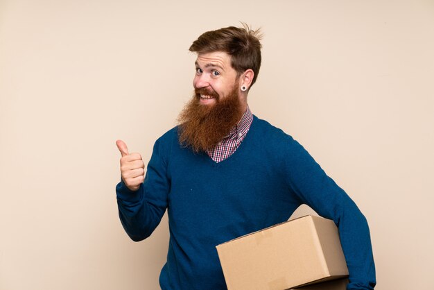 Photo redhead man with long beard over isolated wall holding a box to move it to another site and pointing side