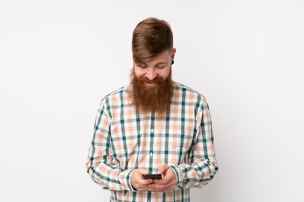 Redhead man with long beard over isolated white wall sending a message with the mobile