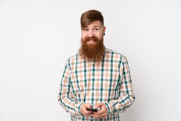 Redhead man with long beard over isolated white wall sending a message with the mobile