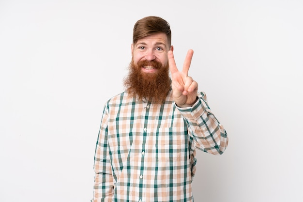 Redhead man with long beard over isolated white wall smiling and showing victory sign