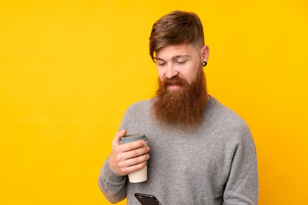 Redhead man with long beard over isolated yellow holding coffee to take away and a mobile