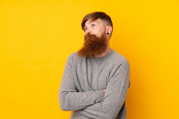 Redhead man with long beard over isolated yellow wall making doubts gesture while lifting the shoulders