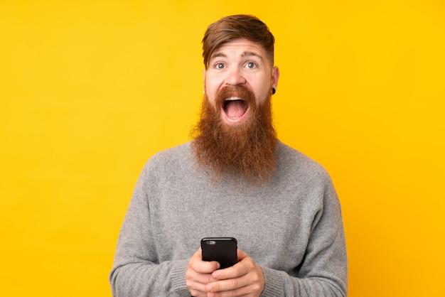 Redhead man with long beard over isolated yellow wall surprised and sending a message