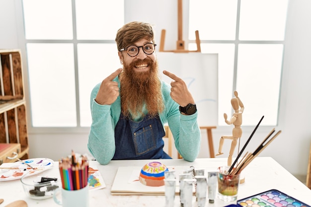 Redhead man with long beard painting clay bowl at art studio smiling cheerful showing and pointing with fingers teeth and mouth. dental health concept.