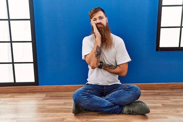 Redhead man with long beard sitting on the floor at empty room thinking looking tired and bored with depression problems with crossed arms.