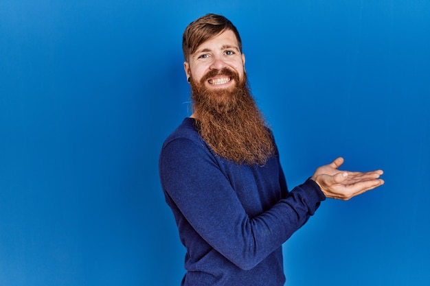 Redhead man with long beard wearing casual blue sweater over blue background pointing aside with hands open palms showing copy space, presenting advertisement smiling excited happy