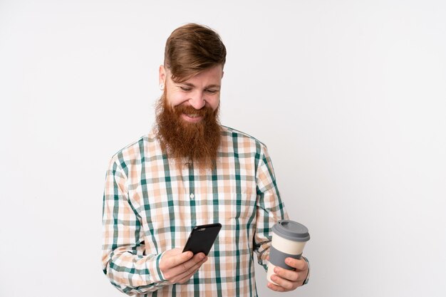 Redhead man with long beard over white wall holding coffee to take away and a mobile