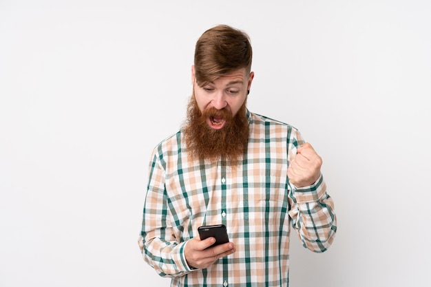 Redhead man with long beard over white wall surprised and sending a message