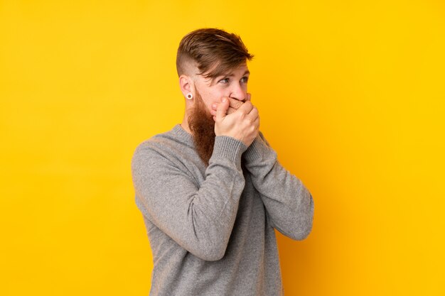 Redhead man with long beard over yellow wall covering mouth and looking to the side