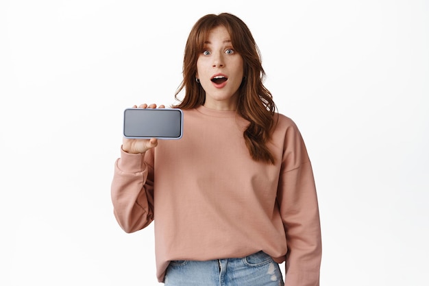 Redhead modern girl showing mobile phone screen, looking surprised, standing against white background.