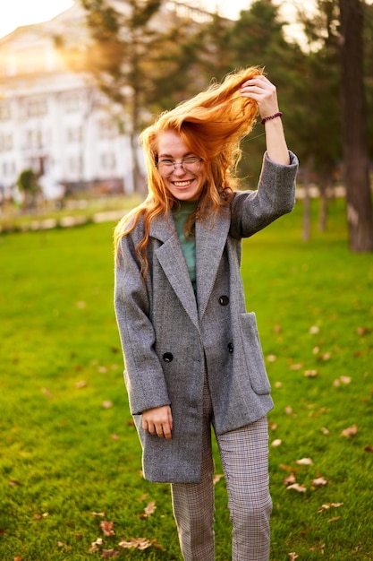 Redhead smiling pretty woman walking in park on sunny autumn day Elegant female poses in stylish authentic outfit has fun laughing on green lawn with vivid foliage leaves Girl enjoys fall season