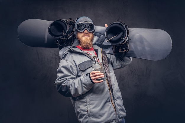 Redhead snowboarder with a full beard in a winter hat and protective glasses dressed in a snowboarding coat holds a snowboard and a bottle of alcohol on his shoulder in a studio. Isolated on gray back