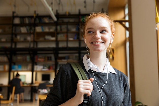 Redhead student posing indoors in library istening music with earphones