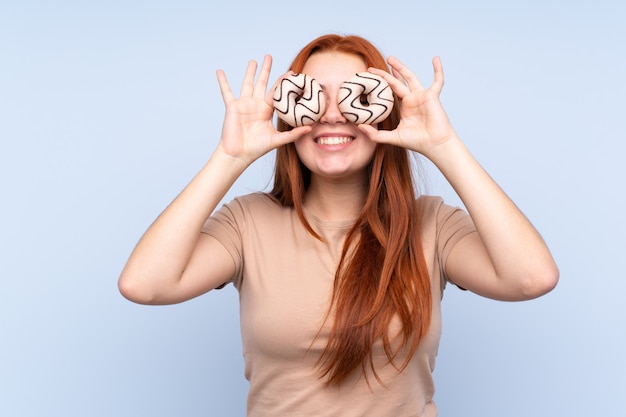 Redhead teenager woman over isolated blue wall holding donuts in eyes