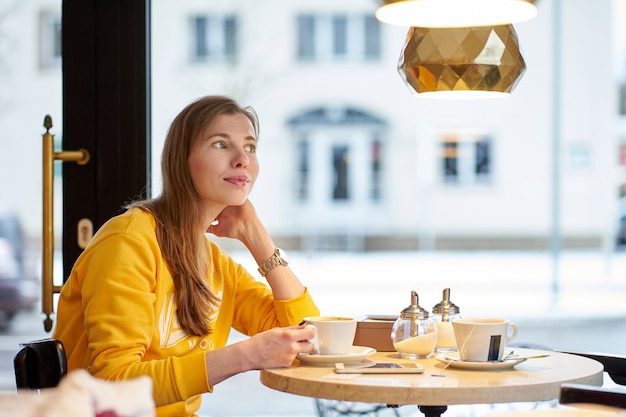 Redhead white girl wearing yellow sweatshirt drinking coffee in cafe and dreaming