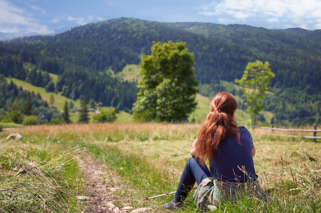 Redhead woman in a dark blue t-shirt sits on the meadow in the mountain area, her face towards the mountains and the fir-tree forest. Girl resting after hiking.