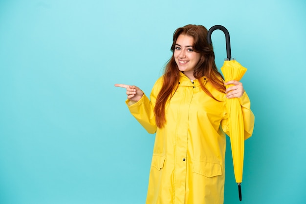 Redhead woman holding an umbrella isolated on blue background pointing finger to the side