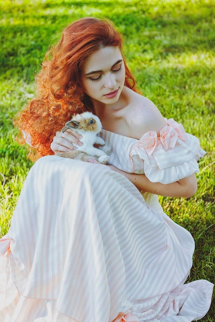 redhead woman in long dress playing with cute fluffy Easter bunny. Easter Day in green spring park.