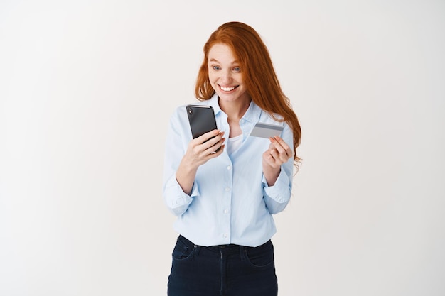 Redhead woman making online order, holding plastic credit card and looking at smartphone screen surprised, white wall