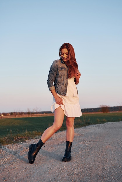 Redhead woman in skirt standing and posing for a camera on the road at evening time