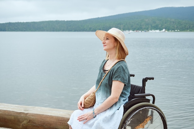 Redhead woman in wheelchair with hat enjoying seascape or lake.