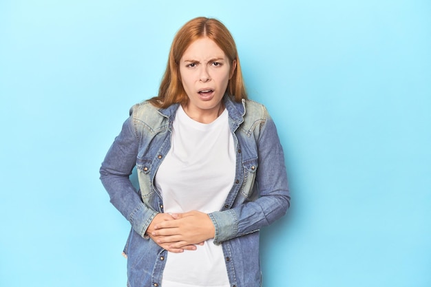 Redhead young woman on blue background having a liver pain stomach ache